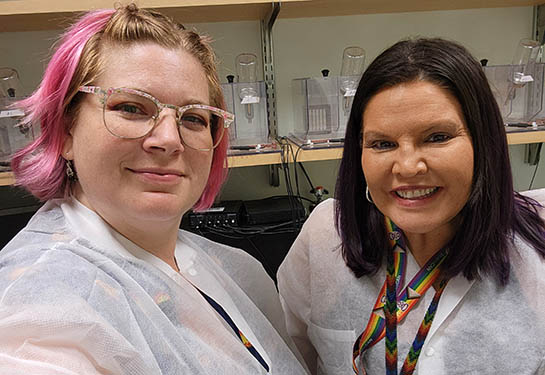 Two researchers in white lab coats stand together in a research facility. 